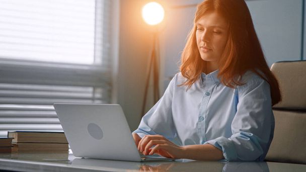 Skoncentrowana pielęgniarka rudowłosa z długimi luźnymi włosami na szarym laptopie komunikująca się z pacjentami online w biurze - Zdjęcie, obraz