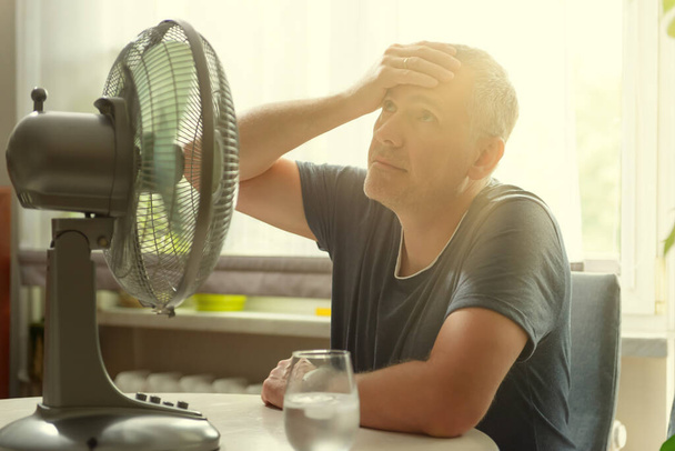 L'homme souffre de chaleur alors qu'il est assis dans la chambre et essaie de se rafraîchir par le ventilateur - Photo, image