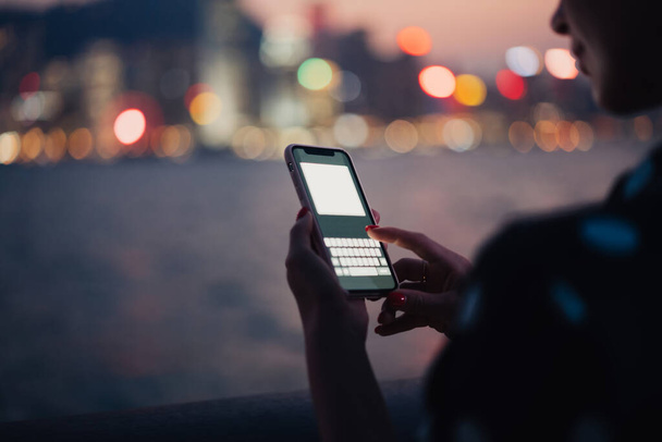 Crop of unrecognizable female pointing finger on screen of modern smartphone in plastic case with messenger mock up on blurred background of night river and bokeh city lights - Φωτογραφία, εικόνα