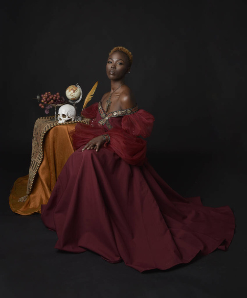 Klassiek portret van een mooie Afrikaanse vrouw in rode renaissance middeleeuwse fantasie jurk, schaduwrijke verlichting op een donkere studio achtergrond. - Foto, afbeelding