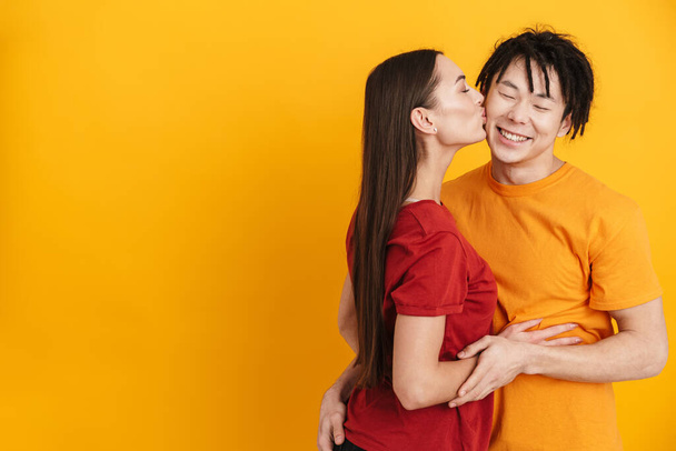 Щаслива багатоетнічна пара в футболках стоїть ізольовано на жовтому тлі, цілується і приймає
 - Фото, зображення