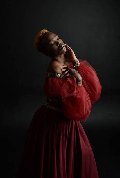 Klasszikus portré csinos afrikai nő visel piros reneszánsz középkori fantasy ruha, árnyékos világítás egy sötét stúdió háttér. - Fotó, kép