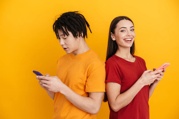 Young multiracial couple posing and using mobile phones isolated over yellow wall - Photo, Image