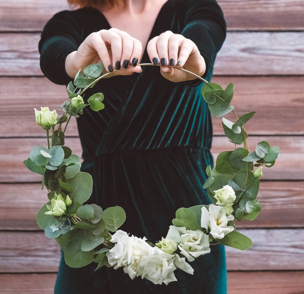 Frauenhände mit einem geschmückten Kranz aus Blumen und Blättern auf einem hölzernen Hintergrund. Kopie, leerer Raum für Text - Foto, Bild