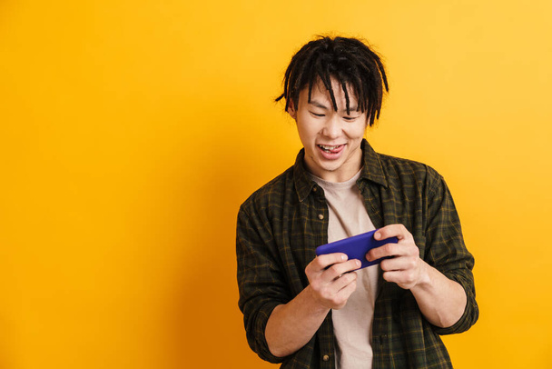 Happy asian man standing over yellow wall background, using mobile phone, playing games - Foto, Bild