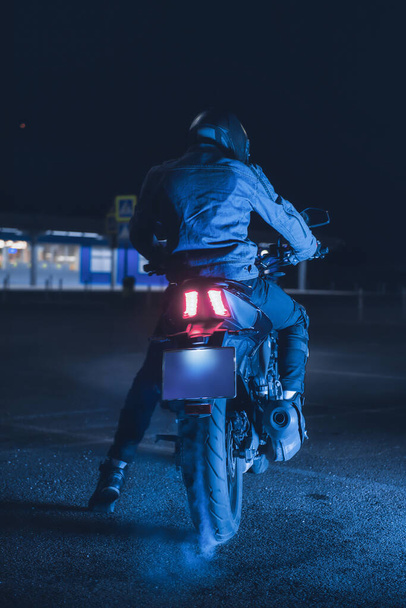 Motorcyclist sits on a motorbike in neon light in an empty parking lot at night - Zdjęcie, obraz