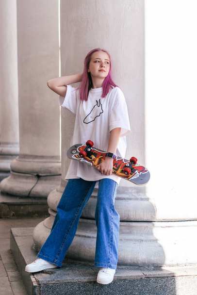 A pink-haired hipster teenage girl in a white T-shirt holding a bright skateboard on a city street on a summer day.Generation Z style,active lifestyle.Copy space. - Фото, зображення