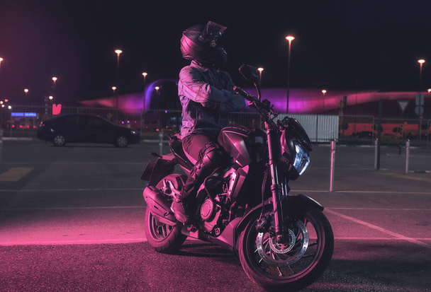 Motorcyclist sits on a motorbike in neon light in an empty parking lot at night - Fotoğraf, Görsel