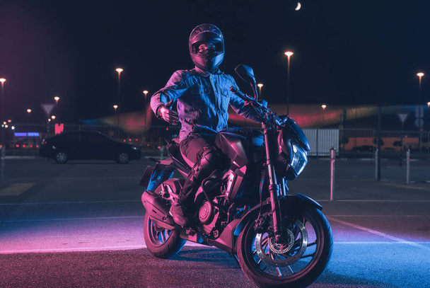 Motorcyclist sits on a motorbike in neon light in an empty parking lot at night - Foto, Bild