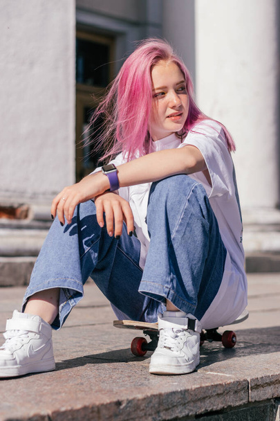 A pink-haired hipster teenage girl in a white T-shirt and jeans is sitting on a skateboard on a city street.Generation Z style,active lifestyle,summer concept.Copy space for text. - Foto, Imagen