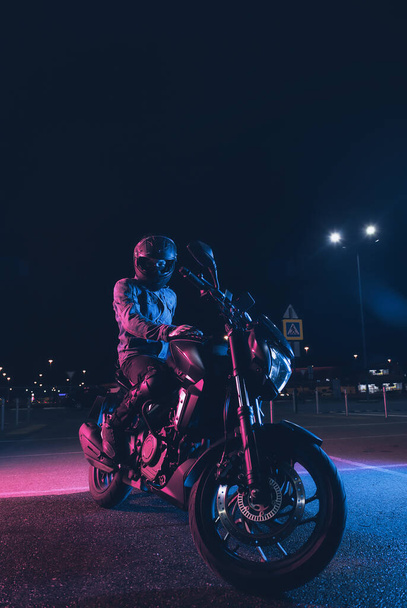 Motorcyclist sits on a motorbike in neon light in an empty parking lot at night - Foto, afbeelding