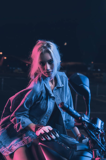Porträt eines Mädchens im Neonlicht auf einem Motorrad in der Nacht auf einem leeren Parkplatz - Foto, Bild