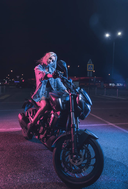 portrait of a girl in the rays of neon light on a motorbike at night in an empty parking lot - Foto, afbeelding