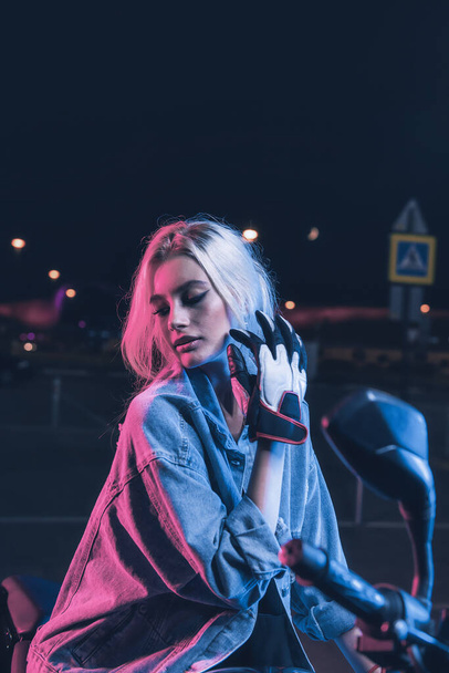 portrait of a girl in the rays of neon light on a motorbike at night in an empty parking lot - Foto, Bild
