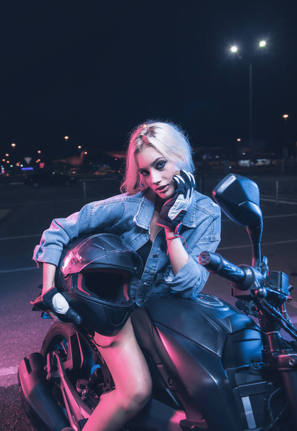 retrato de una chica en los rayos de luz de neón en una moto por la noche en un estacionamiento vacío - Foto, Imagen