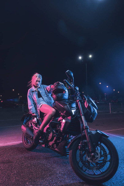 portrait of a girl in the rays of neon light on a motorbike at night in an empty parking lot - Zdjęcie, obraz