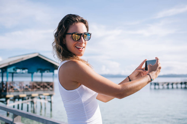 Half length portrait of happy female tourist in sunglasses smiling at camera while using cellular application for shooting live streams in social networks,millennial tourist enjoying vacations in Asia - Foto, Imagen