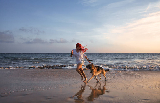 Γυναίκα με μακριά βαμμένα κόκκινα μαλλιά τρέχει με το Γερμανικό Ποιμενικό σκυλί της σε μια παραλία στο ηλιοβασίλεμα με αντανακλάσεις στην υγρή άμμο σε ένα υγιεινό τρόπο ζωής ή διακοπές έννοια. Φιλία, διασκέδαση και απόλαυση στις καλοκαιρινές διακοπές - Φωτογραφία, εικόνα