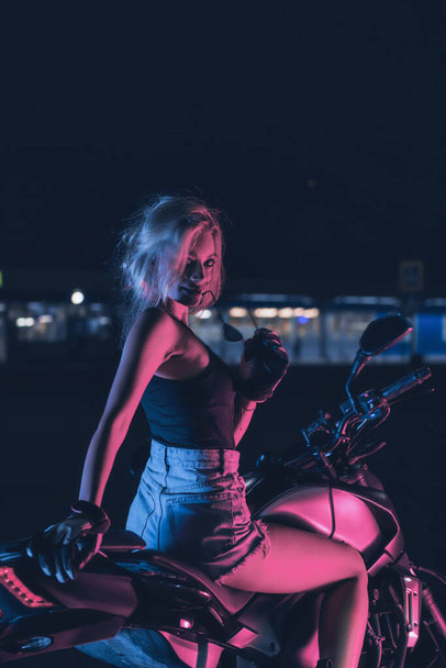 portrait of a girl in the rays of neon light on a motorbike at night in an empty parking lot - Zdjęcie, obraz