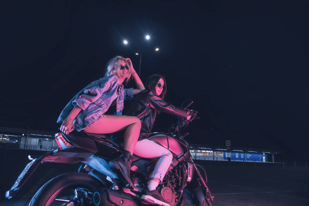 夜の空の駐車場でネオンの光の中でバイクに恋をしている女の子のカップル - 写真・画像