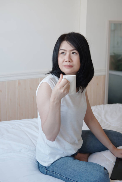 Улыбающаяся азиатка пьет чашку кофе и работает с ноутбуком в спальне во время отпуска. Во время вспышки "Ковод-19" она работает в режиме онлайн, используя технологии из дома. - Фото, изображение