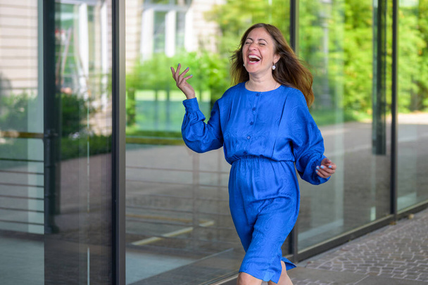Nadšená nadšená žena kráčí šťastně se zářivým úsměvem před skleněnými okny komerční budovy - Fotografie, Obrázek