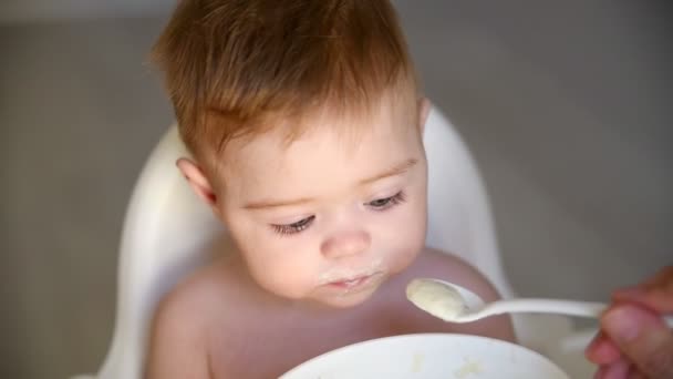 Moeder voedt haar zoontje met pap uit een lepel. Pasgeboren zoontje wordt vies en eet met plezier. De eerste aanvullende voedingsmiddelen voor kinderen. - Video