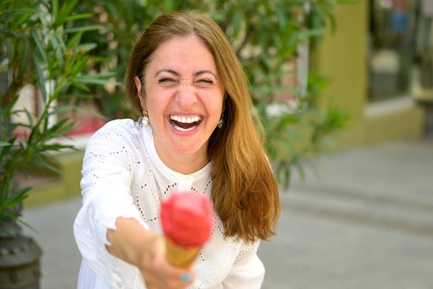 Laughing vivacious woman offering an ice cream cone to camera outdoors on a quiet city street in summer with focus to her face - Photo, Image