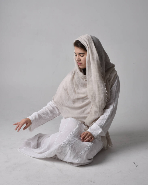 Full length portrait of young woman wearing classical white gown and a head covering veil in biblical style, sitting pose on light studio background. - Fotó, kép