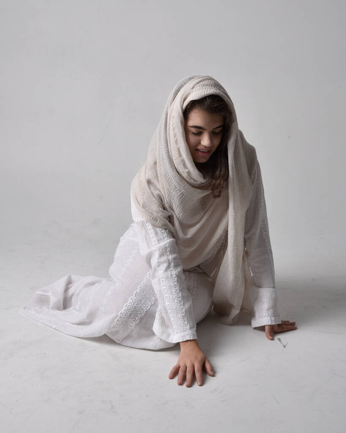 Full length portret van jonge vrouw dragen klassieke witte jurk en een hoofd bekleding sluier in bijbelse stijl, zittende houding op lichte studio achtergrond. - Foto, afbeelding