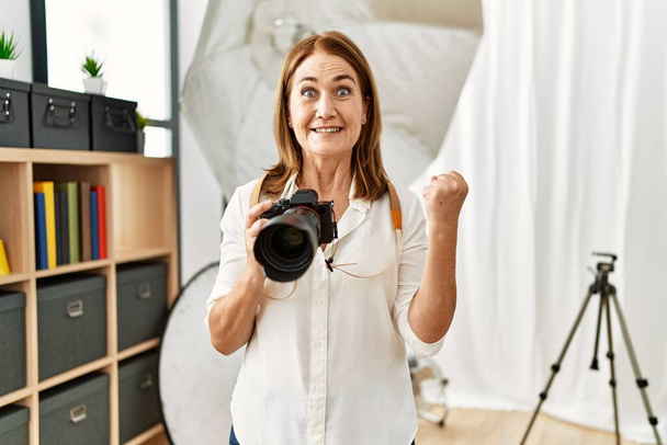 Женщина-фотограф средних лет с фотоаппаратом в фотостудии гордо кричит, празднуя победу и успех, очень взволнована поднятыми руками  - Фото, изображение