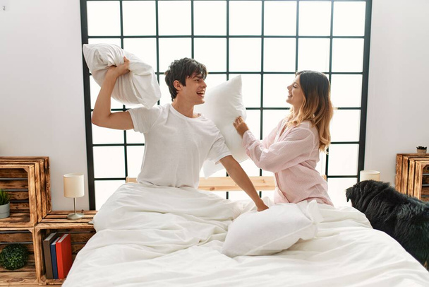 Νεαρό καυκάσιο ζευγάρι χαμογελά ευτυχισμένος αγωνίζονται με μαξιλάρι κάθεται στο κρεβάτι στο σπίτι. - Φωτογραφία, εικόνα