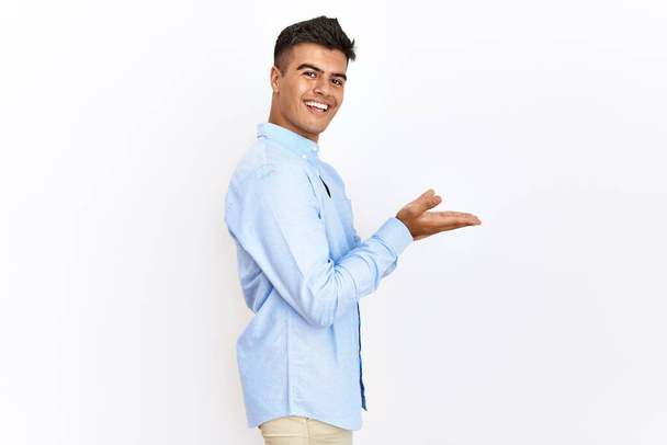 Jeune homme hispanique portant chemise d'affaires debout sur fond isolé pointant de côté avec les mains ouvertes paumes montrant l'espace de copie, présentant la publicité souriant heureux excité  - Photo, image