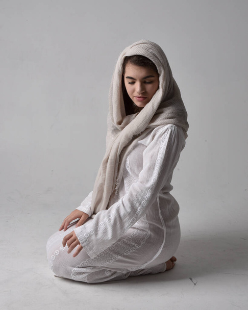 Portrait complet de jeune femme portant une robe blanche classique et un voile couvrant la tête dans un style biblique, posant assise sur un fond de studio léger. - Photo, image