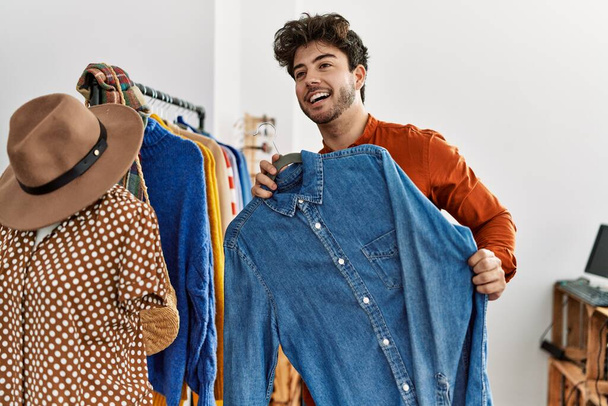 Νεαρός Ισπανός πελάτης χαμογελά χαρούμενος κρατώντας κρεμάστρα με πουκάμισο στο κατάστημα ρούχων. - Φωτογραφία, εικόνα