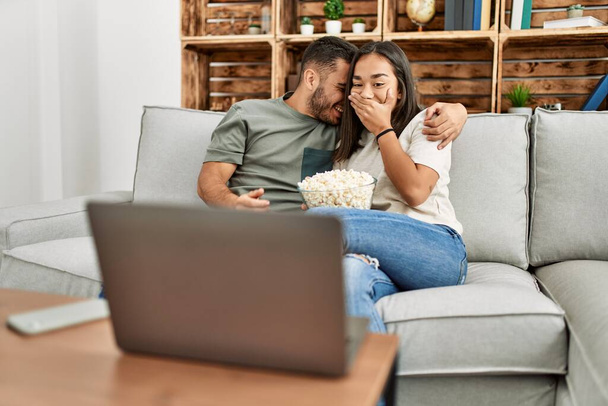 Young hispanic couple watching film and eating popcorn sitting on the sofa at home. - Photo, Image