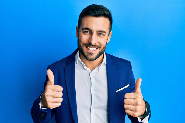 Mladý hispánský obchodník, který má na sobě značku úspěchu obchodní bundy, dělá pozitivní gesto rukou, palce nahoru, usmívá se a je šťastný. veselý výraz a gesto vítěze.  - Fotografie, Obrázek