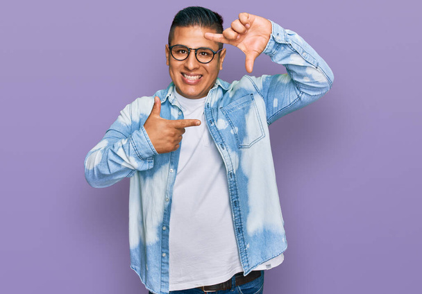 Jovem latino vestindo roupas casuais e óculos sorrindo fazendo moldura com mãos e dedos com rosto feliz. conceito de criatividade e fotografia.  - Foto, Imagem