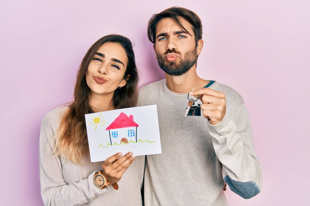 Young hispanic couple holding house draw and key of new home looking at the camera blowing a kiss being lovely and sexy. love expression.  - Φωτογραφία, εικόνα