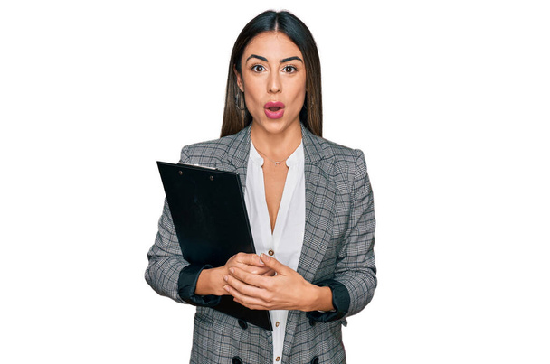 Young hispanic woman wearing business clothes holding clipboard scared and amazed with open mouth for surprise, disbelief face  - Photo, image