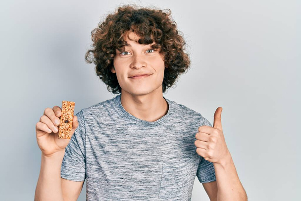 Knappe jongeman die proteïnereep eet als gezonde energie snack glimlachend gelukkig en positief, duim omhoog doen uitstekend en goedkeuring teken  - Foto, afbeelding