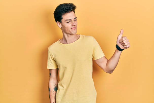 Joven hombre hispano vistiendo camiseta amarilla casual luciendo orgulloso, sonriendo haciendo un gesto hacia un lado  - Foto, Imagen