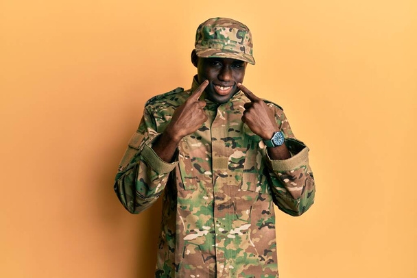 Jovem afro-americano vestindo uniforme do exército sorrindo com a boca aberta, dedos apontando e forçando sorriso alegre  - Foto, Imagem