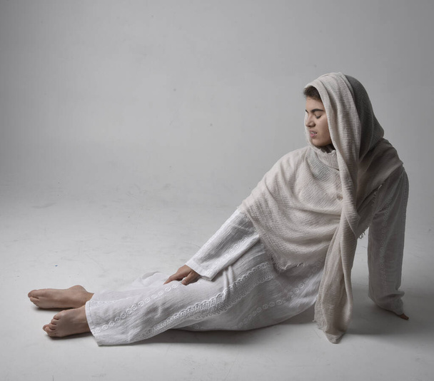 Full length portret van jonge vrouw dragen klassieke witte jurk en een hoofd bekleding sluier in bijbelse stijl, zittende houding op lichte studio achtergrond. - Foto, afbeelding