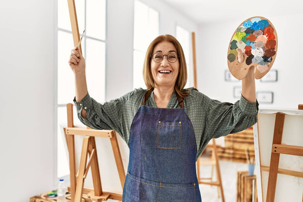 Mulher artista de meia idade sorrindo pintura feliz no estúdio de arte - Foto, Imagem