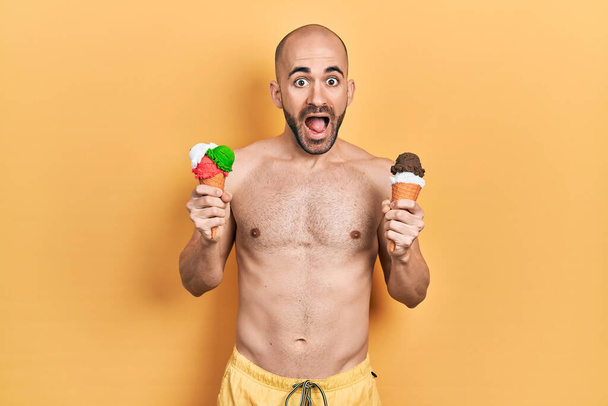 Young bald man wearing swimwear holding two ice cream cones celebrating crazy and amazed for success with open eyes screaming excited.  - Foto, Imagem
