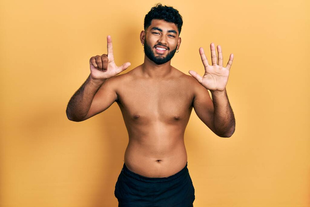 Arabischer Mann mit Bart trägt Badehosen und zeigt mit Finger Nummer sieben nach oben, während er selbstbewusst und glücklich lächelt.  - Foto, Bild