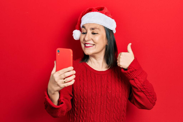 Középkorú spanyol nő visel karácsonyi kalap, amelynek videohívás okostelefon mosolyogva boldog és pozitív, hüvelykujj fel csinál kiváló és jóváhagyási jel  - Fotó, kép