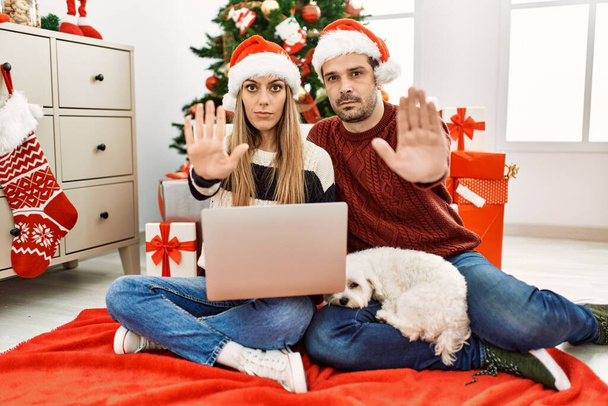 Fiatal pár feleség és férj visel karácsonyi kalap segítségével laptop nyitott kézzel csinál stop jel komoly és magabiztos kifejezés, védelmi gesztus  - Fotó, kép