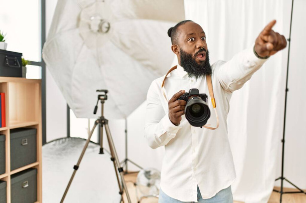 Hombre fotógrafo afroamericano trabajando en estudio de fotografía señalando con el dedo sorprendido delante, boca abierta expresión asombrada, algo en el frente  - Foto, imagen